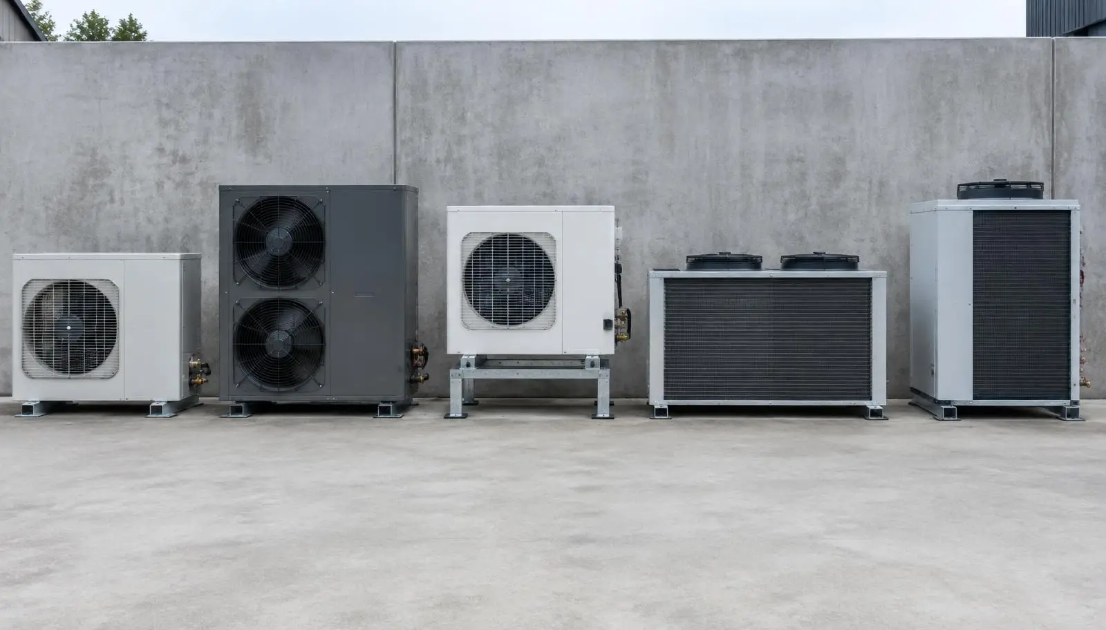 Five outdoor HVAC condenser units of various sizes and designs against a concrete wall.